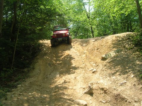 1996 Ford Bronco - A red Jeep Wrangler navigates a challenging dirt incline surrounded by lush green trees.
