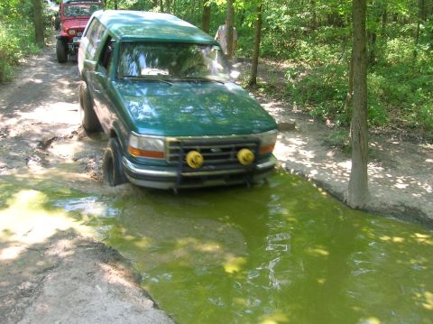 1996 Ford Bronco - A green SUV navigates a muddy trail, splashing through a bright green puddle with another vehicle following.