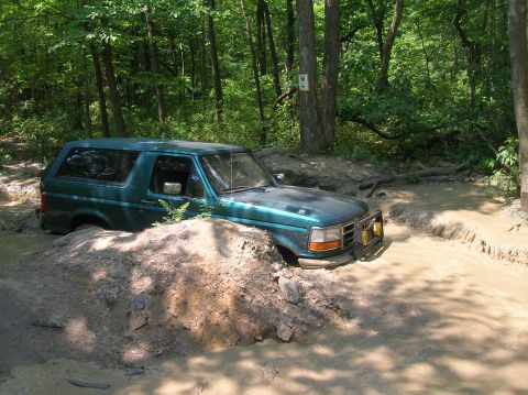 1996 Ford Bronco - A green Ford Bronco navigates a challenging muddy trail, with its front right wheel deep in the mire.