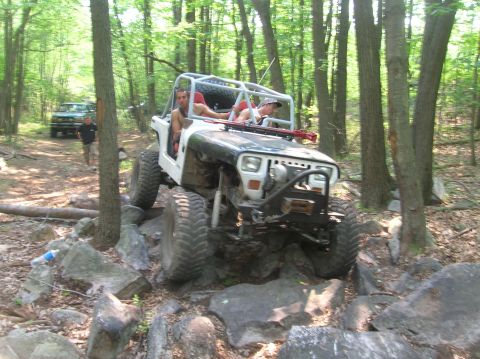 1996 Ford Bronco - A modified Jeep Wrangler tackles a challenging rocky trail in a wooded area.