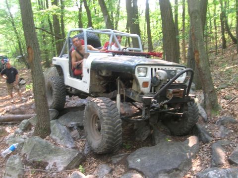 1996 Ford Bronco - A white Jeep Wrangler with a roll cage is driven over large rocks on a wooded trail.