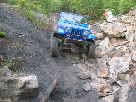 1996 Ford Bronco - A blue Jeep Wrangler tackles a challenging off-road course with steep inclines and rocky terrain.