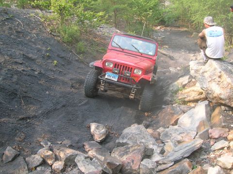 1996 Ford Bronco - A red Jeep Wrangler tackles a challenging off-road course, with a spectator observing from a rocky outcrop.