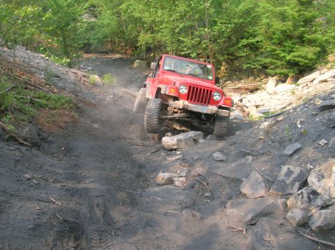 1996 Ford Bronco - A red Jeep Wrangler navigates a challenging off-road trail, kicking up dust as it climbs over rocks.