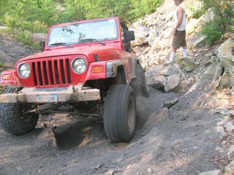1996 Ford Bronco - A red Jeep Wrangler navigates a challenging rocky trail with a person standing on the side.