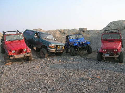 1996 Ford Bronco - Four off-road vehicles are parked on a gravelly surface with rocky hills in the background.