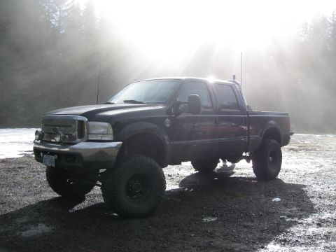 2000 Ford F-250 - A lifted black Ford F-250 pickup truck is parked on a gravel surface with trees and sunbeams in the background.