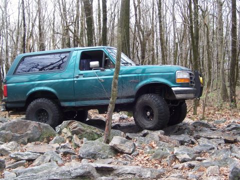 1996 Ford Bronco - A teal Ford Bronco navigates a rocky trail through a forest with bare trees.