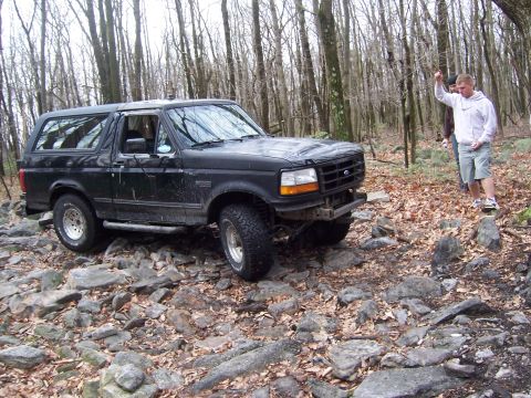 1996 Ford Bronco - A black Ford Bronco navigates a rocky trail in a forest, with a person watching.