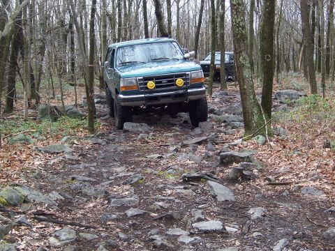 1996 Ford Bronco - A green Ford Bronco navigates a challenging, rocky path through a dense forest with another vehicle following behind.