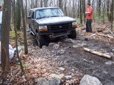 1996 Ford Bronco - A dark-colored Ford Bronco is driven over a rough, rocky path in a wooded area.