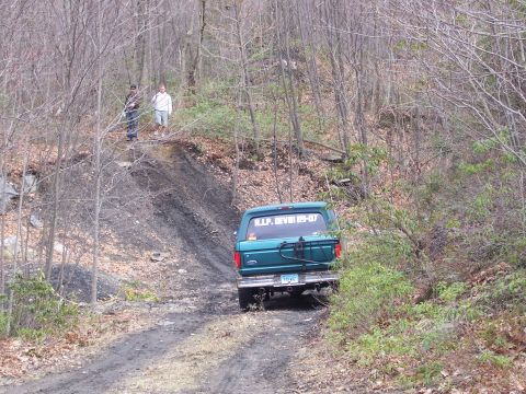 1996 Ford Bronco - A green Ford Bronco drives up a dirt path in a wooded area, with two people standing on a hill above.