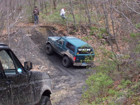 1996 Ford Bronco - A teal Ford Bronco navigates a muddy incline off-road, with another vehicle partially visible in the foreground.