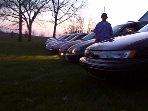 1998 Ford Taurus - A line of sedans is parked on a grassy area at sunset, with a person standing by the front vehicle.