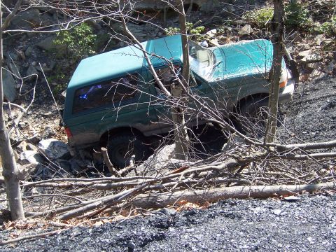 1996 Ford Bronco - A green SUV is partially submerged in debris on a steep, rocky incline, surrounded by trees and branches.