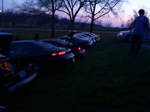 1998 Ford Taurus - Several cars are parked on a grassy area during twilight, with trees in the background.