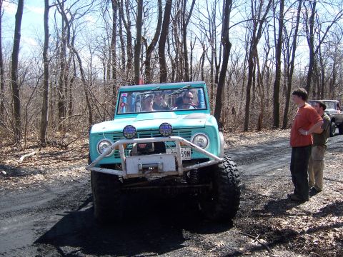 1996 Ford Bronco - A teal first-generation Ford Bronco navigates a wooded area, with two other vehicles visible in the background.