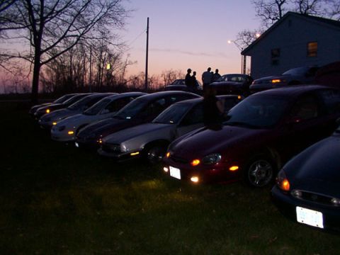 1998 Ford Taurus - A group of cars are parked on a grassy area as the sun sets, with a house and trees visible.