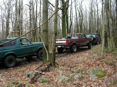 1996 Ford Bronco - A green Ford Bronco is being towed by a maroon Ford F-series pickup truck through a wooded area. Another dark colored SUV is visible behind…