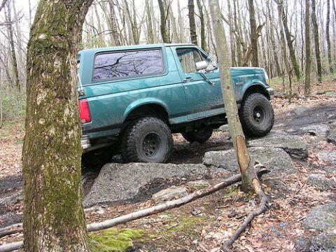1996 Ford Bronco - A teal green Ford Bronco is navigating a rocky, wooded trail. The vehicle is angled to the right, with its front tires on large rocks.