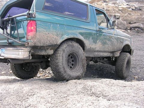 1996 Ford Bronco - The rear driver's side of a muddy, dark green 1996 Ford Bronco is shown in a rocky, outdoor setting. The vehicle has large, off-road tires…