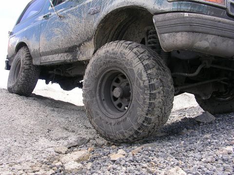 1996 Ford Bronco - The rear passenger side of a muddy, dark green Ford Bronco is shown at a low angle, emphasizing its large, aggressive off-road tires and su…