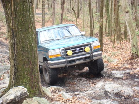 1996 Ford Bronco - A teal Ford Bronco is navigating a rocky, wooded trail. The front of the SUV is angled towards the viewer, with its tires positioned on lar…