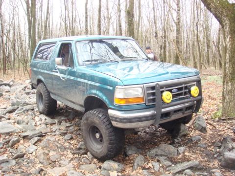 1996 Ford Bronco - A teal 1996 Ford Bronco is shown off-road on a rocky trail surrounded by trees. The vehicle has a brush guard with two smiley face fog ligh…