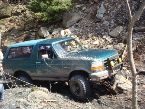 1996 Ford Bronco - A teal Ford Bronco is shown navigating a rocky, off-road trail. The vehicle is covered in mud, indicating it is actively being used for off…