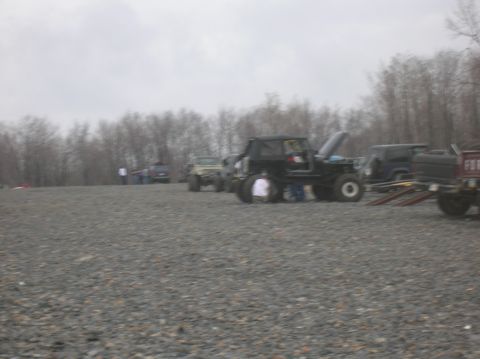 1996 Ford Bronco - A group of modified off-road vehicles, including a black Jeep with its hood open, are parked on a gravel lot with trees in the background.