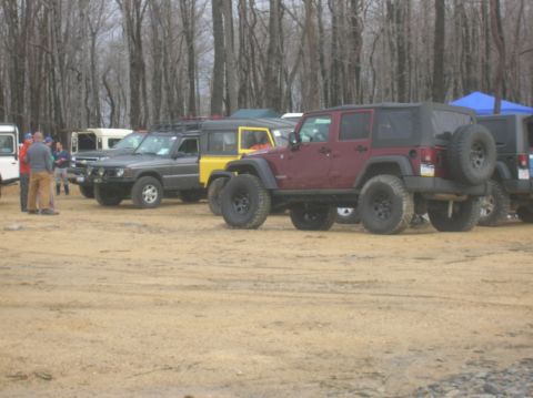 1996 Ford Bronco - A group of off-road vehicles are parked on a dirt lot with trees in the background. A maroon Jeep Wrangler is prominently featured in the f…