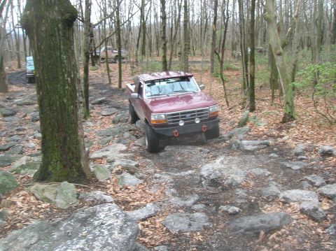 1996 Ford Bronco - A maroon Ford F-150 pickup truck navigates a rocky, wooded trail. The truck is angled towards the viewer, with its front bumper and headlig…