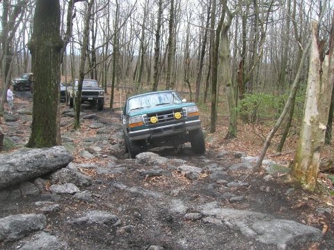 1996 Ford Bronco - A green Ford Bronco is navigating a rocky, wooded trail. The vehicle is angled towards the camera, showing its front grille and headlights…