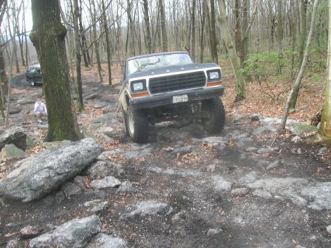 1996 Ford Bronco - A lifted Ford Bronco is navigating a rocky, wooded trail. A person is sitting on rocks to the left of the trail.