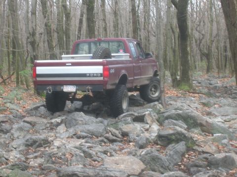 1996 Ford Bronco - A lifted, maroon Ford pickup truck is navigating a rocky, wooded trail. The truck has large off-road tires, a toolbox in the bed, and a spa…