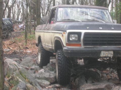 1996 Ford Bronco - A lifted, two-tone Ford truck navigates a rocky, wooded trail. The front of the truck is visible, showing its grille, headlights, and a lic…