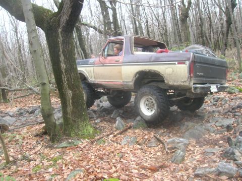 1996 Ford Bronco - A lifted, two-tone brown and tan Ford Bronco is navigating a rocky, wooded trail. The vehicle has large off-road tires and appears to be a…
