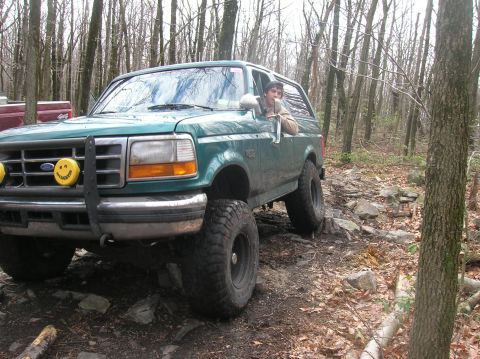 1996 Ford Bronco - A green Ford Bronco is navigating a rocky, wooded trail. The driver is visible through the open driver's side window.