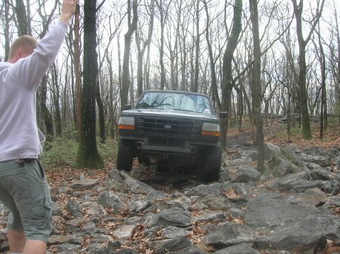 1996 Ford Bronco - A black Ford Bronco is navigating a rocky, wooded trail. A person in a light-colored hoodie and green shorts is visible in the foreground,…