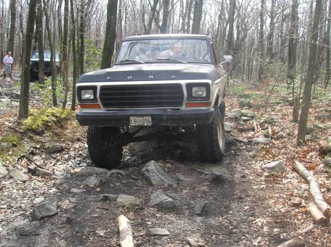 1996 Ford Bronco - A lifted, dark-colored Ford Bronco is driving on a rocky, wooded trail. The front of the vehicle is facing the camera, showing its grille,…