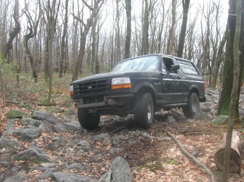 1996 Ford Bronco - A black Ford Bronco is shown from a 3/4 front angle, navigating a rocky, wooded trail.