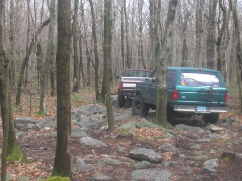 1996 Ford Bronco - Two off-road vehicles, a green SUV and a red pickup truck, are parked on a rocky, wooded trail. The SUV is a Ford Bronco with a visible lic…