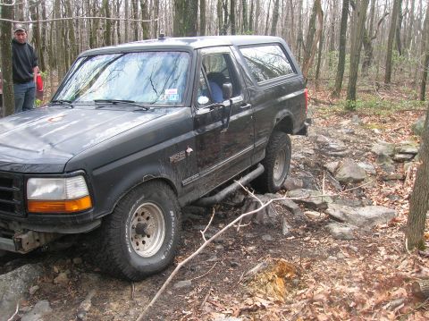 1996 Ford Bronco - A black 1990s Ford Bronco is shown navigating a rocky, wooded trail. The front driver's side of the vehicle is prominently featured, with i…