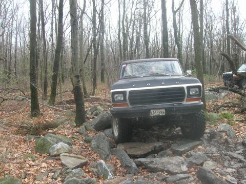 1996 Ford Bronco - A black Ford Bronco is navigating a rocky, wooded trail. The vehicle is positioned at an angle, with its front grille and headlights clearl…