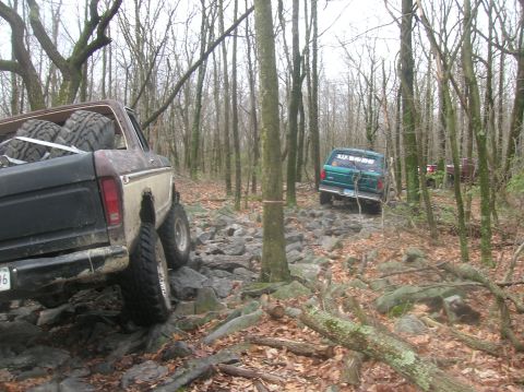1996 Ford Bronco - Two off-road vehicles navigate a rocky, wooded trail. The foreground shows the rear of a lifted pickup truck with large tires, while a gree…