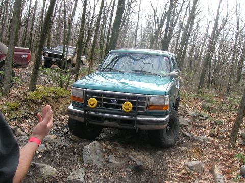 1996 Ford Bronco - A teal Ford Bronco is driving on a rocky, wooded trail. The front of the vehicle is visible, featuring yellow fog lights with smiley faces…