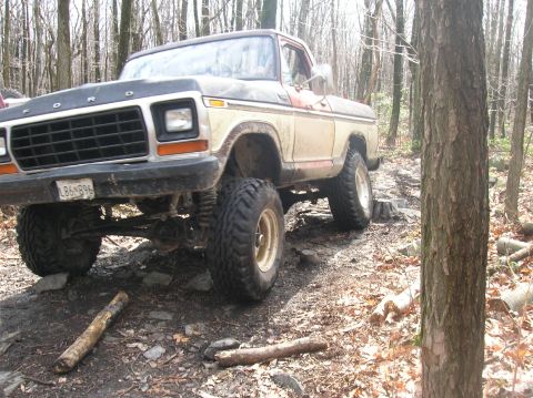 1996 Ford Bronco - A lifted, older model Ford pickup truck is navigating a rocky, wooded trail. The truck is dirty and appears to be off-roading.
