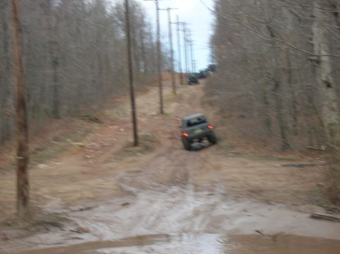 1996 Ford Bronco - A dark-colored SUV drives down a muddy, wooded trail lined with utility poles. The vehicle is angled away from the camera and appears to be…