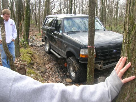 1996 Ford Bronco - A black Ford Bronco is parked on a wooded trail. A person in a grey hoodie is visible in the foreground, and another person in a white hood…