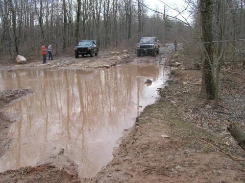1996 Ford Bronco - Two Ford Broncos navigate a muddy trail with a large puddle in the foreground. The vehicles are surrounded by a dense forest.